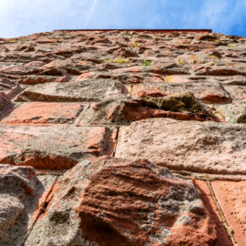 Das Bild zeigt eine Nahaufnahme einer Steinmauer, die aus großen, unregelmäßig geformten Steinen in verschiedenen Rot- und Grautönen besteht. Zwischen einigen Steinen wachsen kleine Pflanzen, und im Hintergrund ist ein klarer blauer Himmel mit wenigen Schleierwolken zu sehen.
