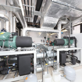 Das Bild zeigt einen Technikraum mit verschiedenen Industrieanlagen, darunter große Rohre, Leitungen und Maschinen. Zwei große grüne Geräte mit schwarzen zylindrischen Komponenten sind auf weißen Gestellen montiert. An Decke und Wänden verlaufen zahlreiche metallische Rohrleitungen, ergänzt durch Steuerungselemente und Messanzeigen.