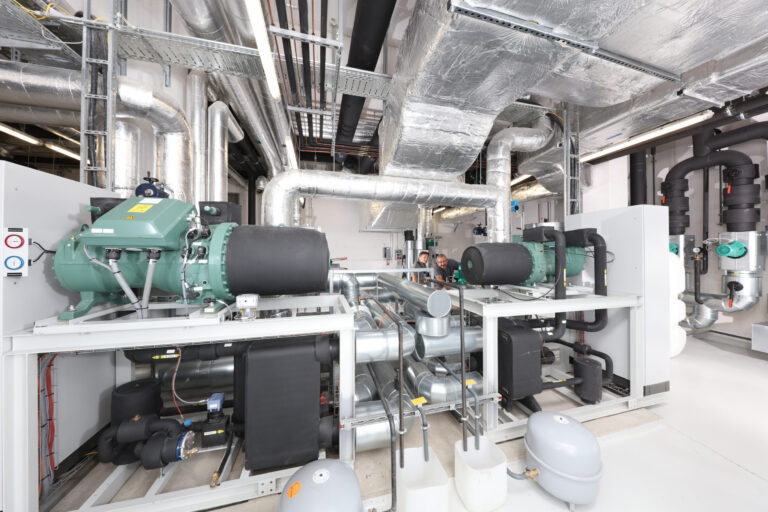 Das Bild zeigt einen Technikraum mit verschiedenen Industrieanlagen, darunter große Rohre, Leitungen und Maschinen. Zwei große grüne Geräte mit schwarzen zylindrischen Komponenten sind auf weißen Gestellen montiert. An Decke und Wänden verlaufen zahlreiche metallische Rohrleitungen, ergänzt durch Steuerungselemente und Messanzeigen.