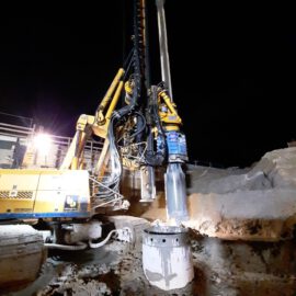 Das Bild zeigt eine nächtliche Baustelle mit einer großen gelben Bohrmaschine im Einsatz. Die Maschine verfügt über mehrere hydraulische Arme und Schläuche und bohrt in den Boden, was an einem zylindrischen Loch und umliegendem Bohrmaterial erkennbar ist. Eine starke Lichtquelle beleuchtet die Szene, im Hintergrund sind weitere Strukturen sichtbar.