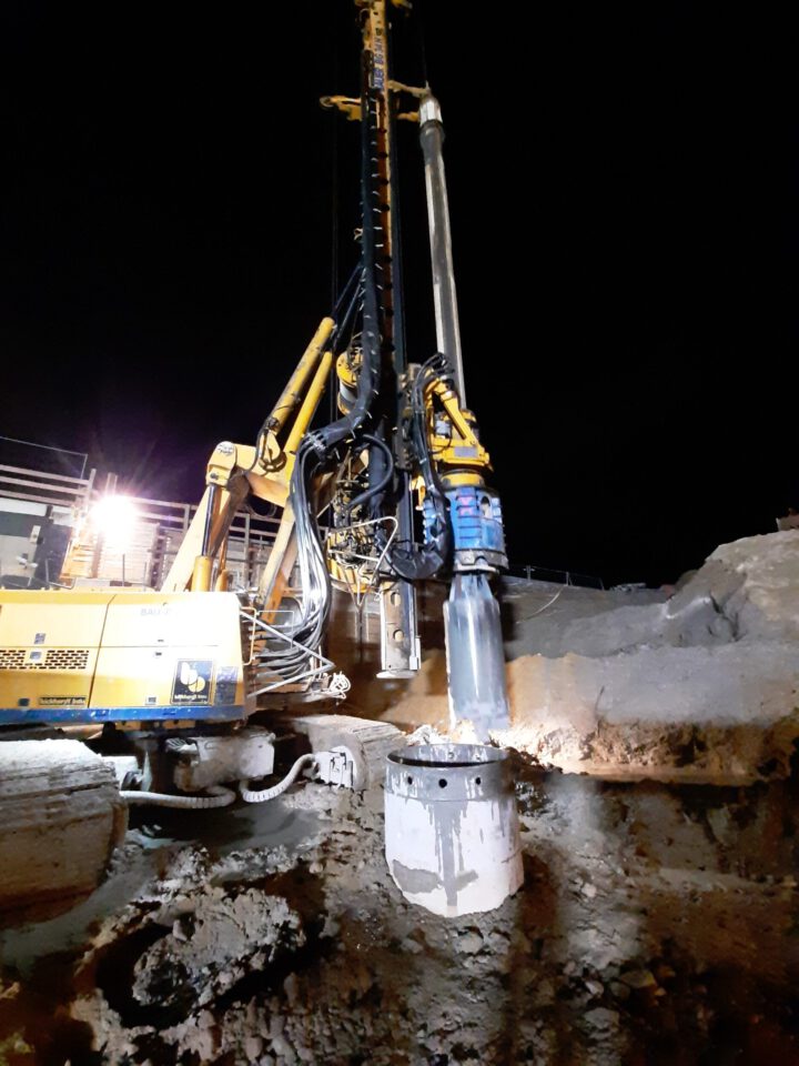 Das Bild zeigt eine nächtliche Baustelle mit einer großen gelben Bohrmaschine im Einsatz. Die Maschine verfügt über mehrere hydraulische Arme und Schläuche und bohrt in den Boden, was an einem zylindrischen Loch und umliegendem Bohrmaterial erkennbar ist. Eine starke Lichtquelle beleuchtet die Szene, im Hintergrund sind weitere Strukturen sichtbar.