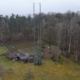 Das Bild zeigt ein Außengelände mit einem hohen Metallturm in der Mitte, umgeben von einer Wiese und einem Waldgebiet. Ein eingezäunter Bereich enthält technische Anlagen oder Baumaterialien. Im Hintergrund sind weitere Bäume und teilweise sichtbare Gebäude zu erkennen, bei bewölktem Himmel.