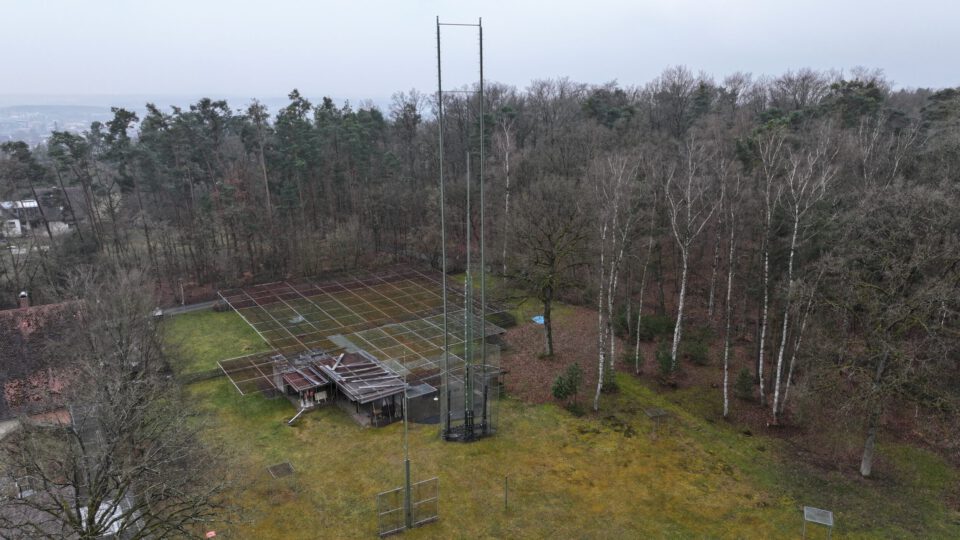 Das Bild zeigt ein Außengelände mit einem hohen Metallturm in der Mitte, umgeben von einer Wiese und einem Waldgebiet. Ein eingezäunter Bereich enthält technische Anlagen oder Baumaterialien. Im Hintergrund sind weitere Bäume und teilweise sichtbare Gebäude zu erkennen, bei bewölktem Himmel.