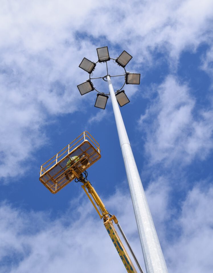 Das Bild zeigt einen hohen Lichtmast mit mehreren Flutlichtstrahlern, die kreisförmig am oberen Ende angeordnet sind. Ein gelber Hubsteiger mit einem Arbeiter im Korb ist bis zur Spitze des Masts ausgefahren. Im Hintergrund ist ein blauer Himmel mit vereinzelten Wolken zu sehen.