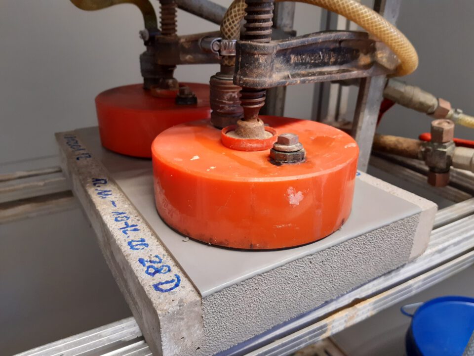 Das Bild zeigt einen Versuchsaufbau in einem Labor mit zwei orangefarbenen, runden Gewichten oder Druckelementen auf einem grauen rechteckigen Block. Der Block besteht vermutlich aus Beton oder einem ähnlichen Material. Metallklemmen und Schläuche deuten auf eine technische Prüfung der Materialeigenschaften hin.