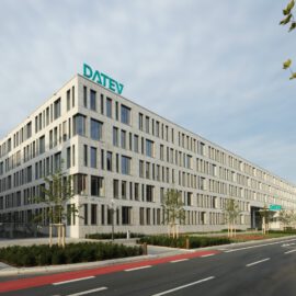 Das Bild zeigt ein großes, modernes Bürogebäude mit dem Schriftzug „DATEV“ auf dem Dach. Vor dem Gebäude verläuft eine Straße mit Fahrradweg, daneben befinden sich Bäume und Grünflächen. Rechts im Bild sind zwei weiße Lastwagen zu sehen, der Himmel ist teilweise bewölkt.