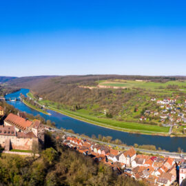 Das Bild zeigt eine Luftaufnahme einer malerischen Landschaft mit einem Fluss, der sich durch ein Tal schlängelt. Links vom Fluss liegt eine historische Burg mit roten Ziegeldächern und Steinmauern, umgeben von Bäumen und Grünflächen. Daneben befindet sich eine kleine Stadt mit zahlreichen Häusern, ebenfalls mit roten Dächern.