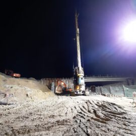 Das Bild zeigt eine nächtliche Baustelle mit einer großen Bohrmaschine im Zentrum. Der Boden ist von Erde und Reifenspuren bedeckt, links liegen mehrere Haufen aus Sand oder Erdmaterial. Im Hintergrund ist ein teilweise errichtetes Bauwerk zu sehen, möglicherweise eine Brücke oder ein Überbau, umgeben von weiteren Baumaschinen und Materialien.