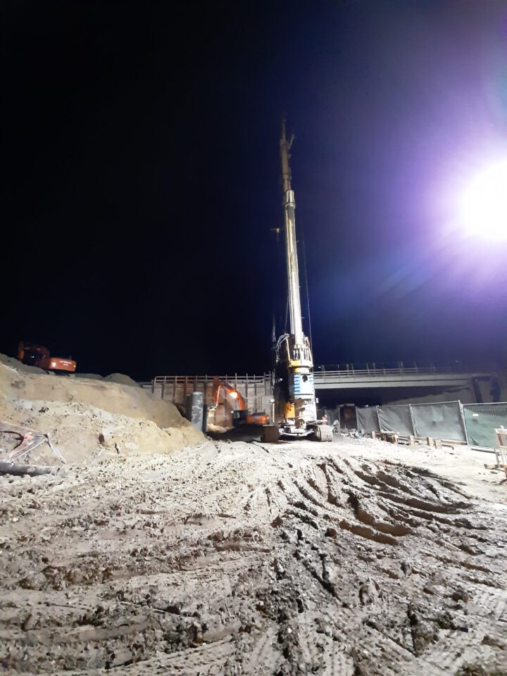 Das Bild zeigt eine nächtliche Baustelle mit einer großen Bohrmaschine im Zentrum. Der Boden ist von Erde und Reifenspuren bedeckt, links liegen mehrere Haufen aus Sand oder Erdmaterial. Im Hintergrund ist ein teilweise errichtetes Bauwerk zu sehen, möglicherweise eine Brücke oder ein Überbau, umgeben von weiteren Baumaschinen und Materialien.
