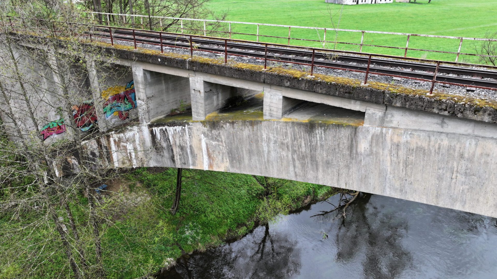 Das Bild zeigt eine zweistöckige Betonbrücke, wobei die obere Ebene mit Bahngleisen und einem Metallgeländer ausgestattet ist. Die untere Ebene ist eine Bogenbrücke, die über einen Fluss führt. Umgeben ist die Brücke von grüner Wiese und Bäumen.