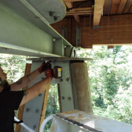 Das Bild zeigt eine Person mit Gehörschutz und Handschuhen, die mit einem Elektrowerkzeug an einer großen Metallkonstruktion arbeitet. Die Person steht auf einer Plattform im Freien, umgeben von Bäumen. Die Konstruktion besteht aus Metall und möglicherweise auch Holzbalken.