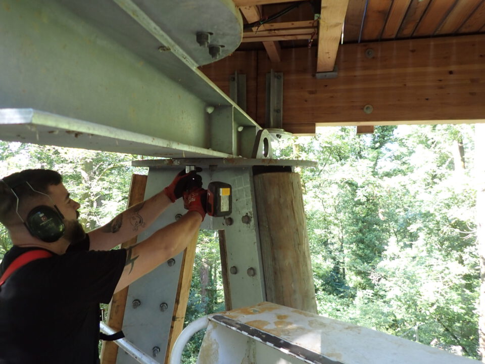 Das Bild zeigt eine Person mit Gehörschutz und Handschuhen, die mit einem Elektrowerkzeug an einer großen Metallkonstruktion arbeitet. Die Person steht auf einer Plattform im Freien, umgeben von Bäumen. Die Konstruktion besteht aus Metall und möglicherweise auch Holzbalken.