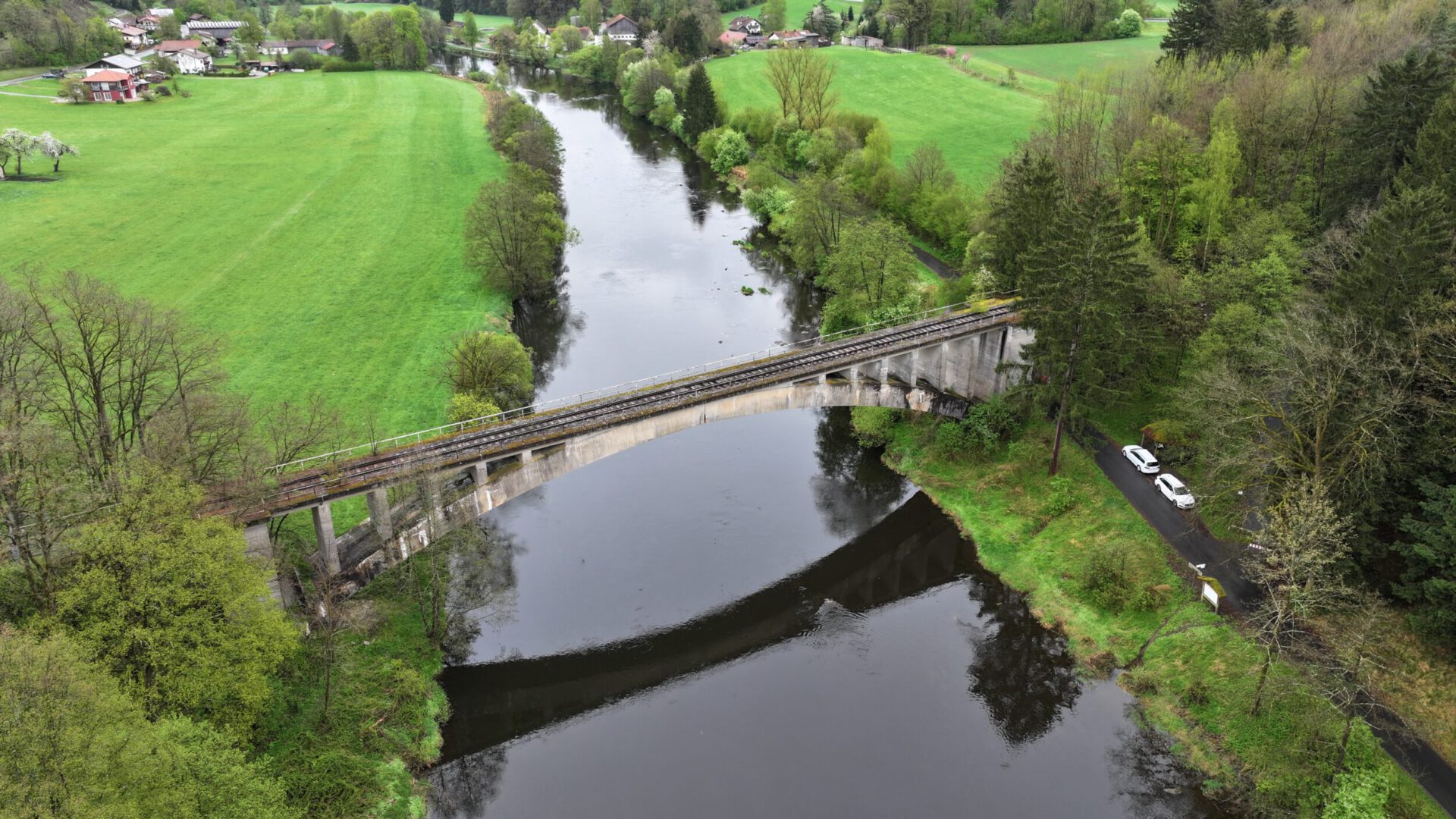 Das Bild zeigt eine idyllische Landschaft mit einem Fluss, der durch die Mitte verläuft, und einer alten Steinbrücke, die vermutlich für den Bahnverkehr genutzt wird. Umgeben ist die Szene von grünen Feldern, Bäumen und einigen Häusern in der Ferne. Am rechten Bildrand verläuft eine Straße mit zwei geparkten Autos nahe eines Waldgebiets.