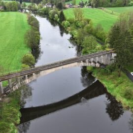 Das Bild zeigt eine idyllische Landschaft mit einem Fluss, der durch die Mitte verläuft, und einer alten Steinbrücke, die vermutlich für den Bahnverkehr genutzt wird. Umgeben ist die Szene von grünen Feldern, Bäumen und einigen Häusern in der Ferne. Am rechten Bildrand verläuft eine Straße mit zwei geparkten Autos nahe eines Waldgebiets.
