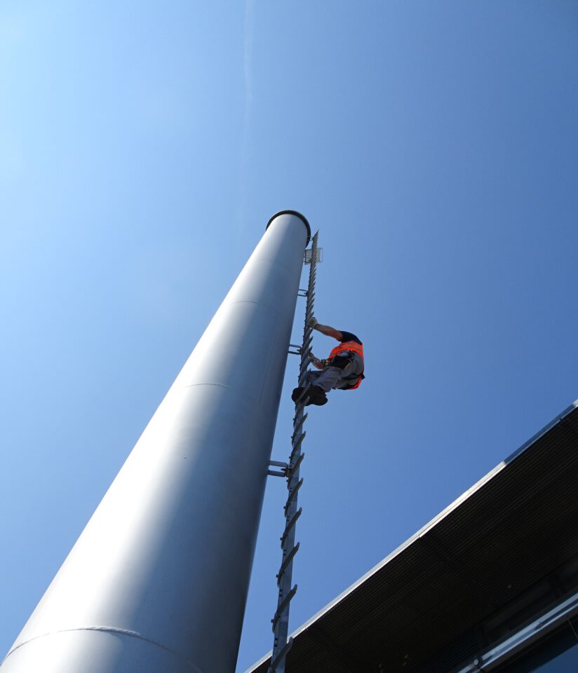 Das Bild zeigt eine Person mit Sicherheitsgurt und orangefarbener Weste, die eine hohe Metallleiter an einem senkrechten Bauwerk hinaufklettert. Im Hintergrund ist ein klarer blauer Himmel zu sehen, und am rechten Bildrand ist ein Teil eines Gebäudedachs erkennbar.