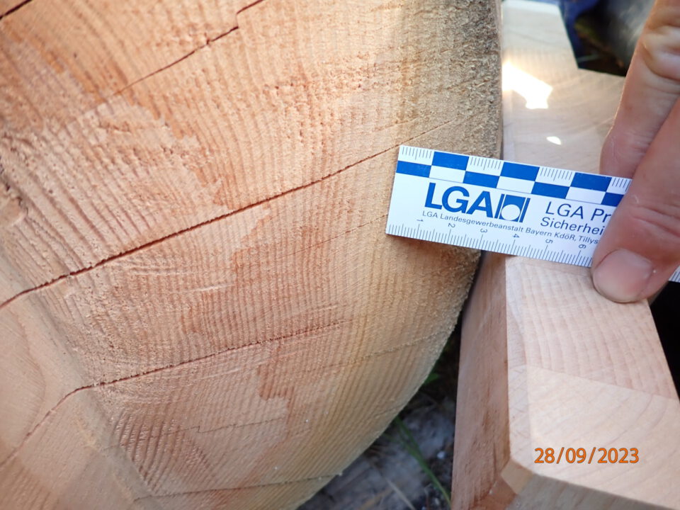 Das Bild zeigt eine Nahaufnahme eines Holzstücks mit sichtbaren Maserungen und Rissen. Eine Hand hält ein Lineal mit der Aufschrift LGA vor das Holz, um die Struktur zu vermessen. Unten rechts ist ein Datum eingeblendet.