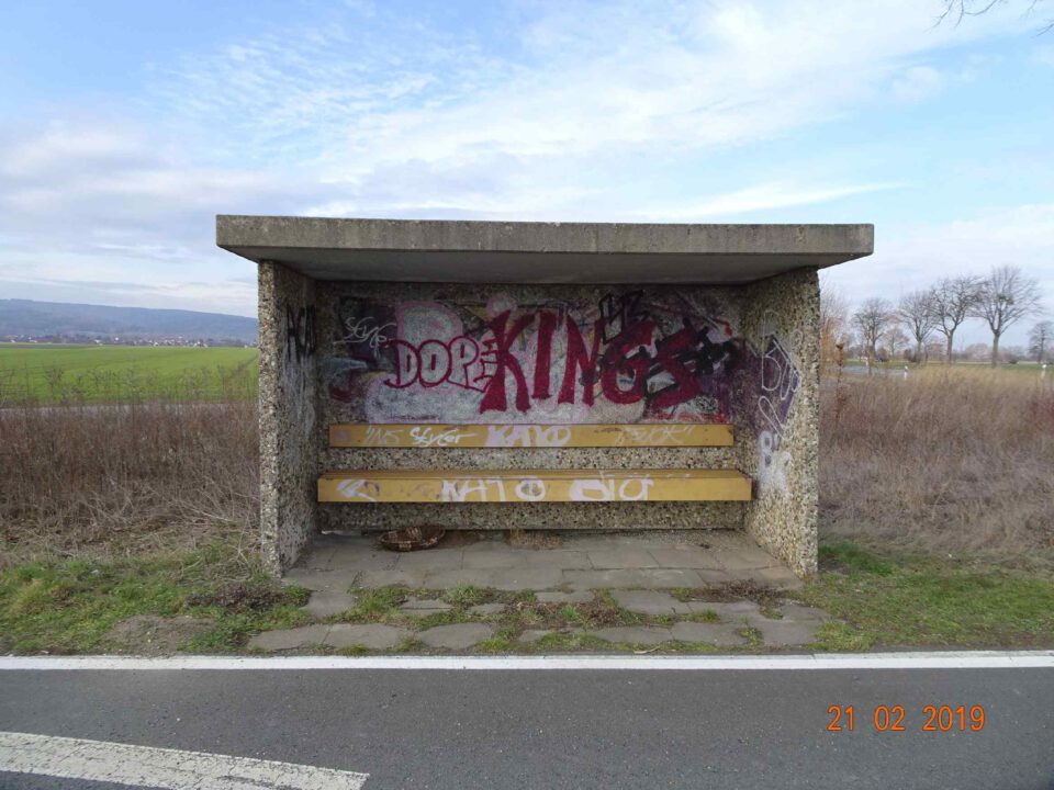 Das Bild zeigt eine kleine Bushaltestelle aus Beton mit Sitzbänken und Graffiti an der Rückwand. Die Haltestelle befindet sich an einer Straße, im Hintergrund sind eine Wiese und Bäume zu sehen. Das Umfeld wirkt ländlich und ruhig.