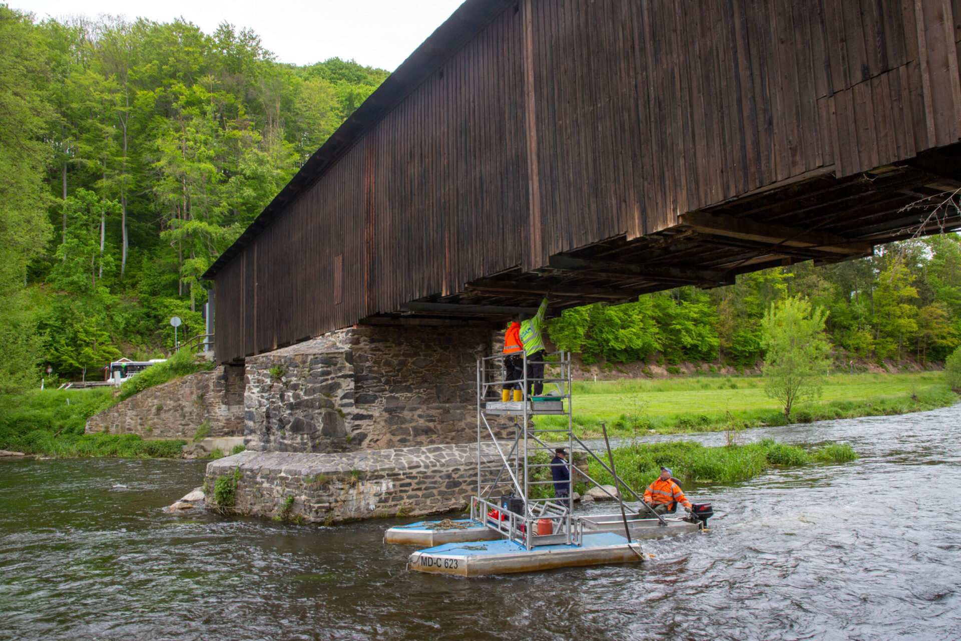 Das Bild zeigt eine überdachte Holzbrücke, die über einen Fluss führt. Unter der Brücke befinden sich zwei Arbeiter in orangefarbener Sicherheitskleidung auf einer schwimmenden Plattform, einer steht auf einem Gerüst, der andere sitzt am Rand der Plattform. Die Brücke ruht auf steinernen Pfeilern, im Hintergrund ist dichte grüne Vegetation zu sehen.