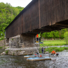 Das Bild zeigt eine überdachte Holzbrücke, die über einen Fluss führt. Unter der Brücke befinden sich zwei Arbeiter in orangefarbener Sicherheitskleidung auf einer schwimmenden Plattform, einer steht auf einem Gerüst, der andere sitzt am Rand der Plattform. Die Brücke ruht auf steinernen Pfeilern, im Hintergrund ist dichte grüne Vegetation zu sehen.