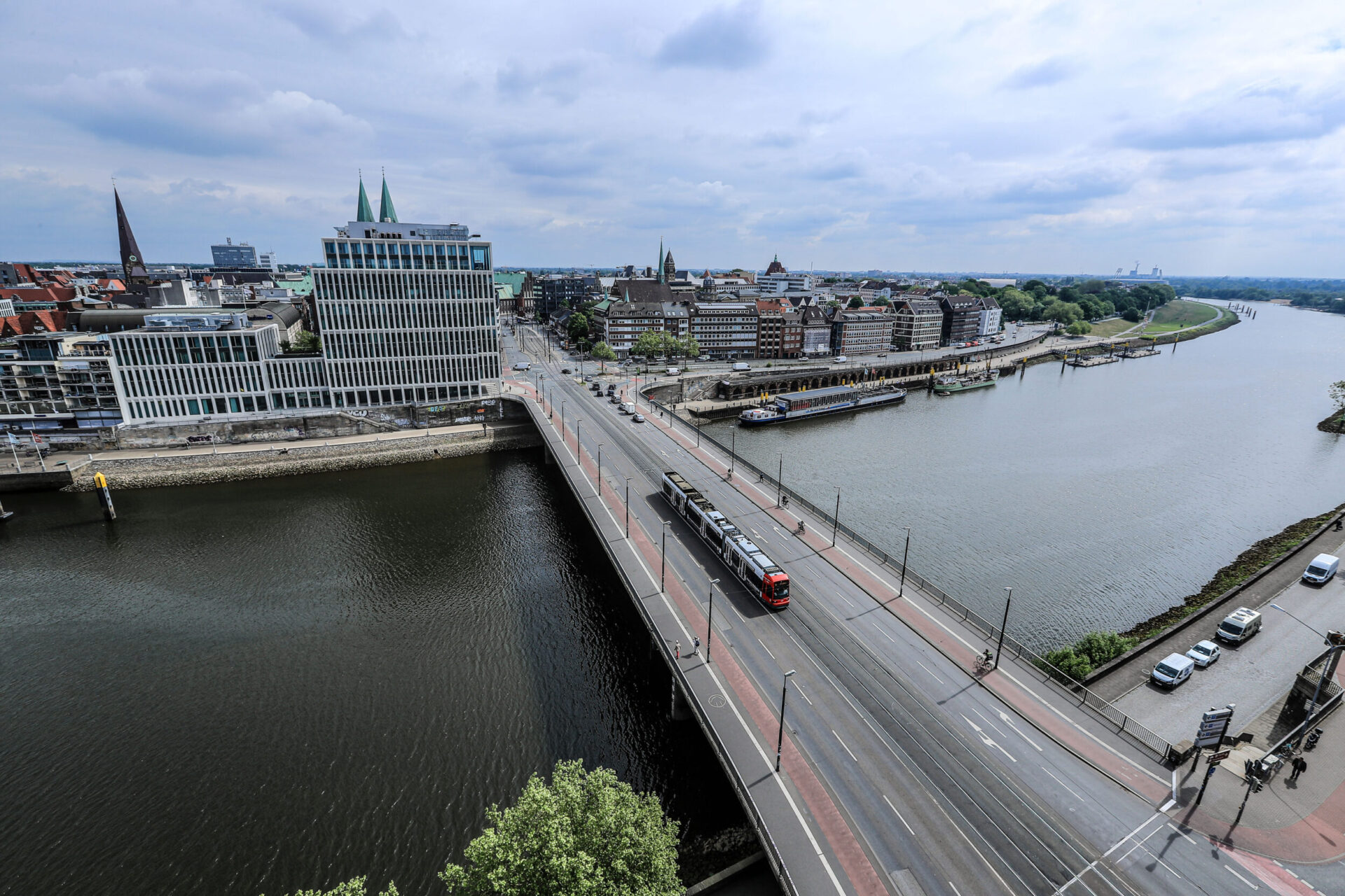 Das Bild zeigt eine Luftaufnahme einer Stadt mit einem Fluss, der sich durch das Stadtgebiet zieht. Eine Brücke überquert den Fluss, auf der eine Straßenbahn oder ein Zug unterwegs ist. Die Stadt vereint moderne Gebäude mit grünen Türmen und ältere Architektur, der Himmel ist teilweise bewölkt.