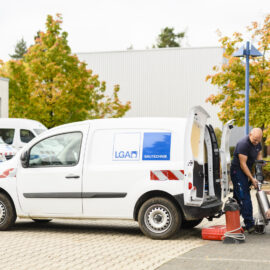 Das Bild zeigt zwei Personen bei der Arbeit neben einem weißen Transporter mit der Aufschrift „LGA BAUTECHNIK“. Die Hecktüren des Fahrzeugs sind geöffnet, und es sind verschiedene Geräte im und um den Wagen zu sehen. Im Hintergrund stehen weitere Fahrzeuge und Gebäude, umgeben von herbstlich gefärbten Bäumen.