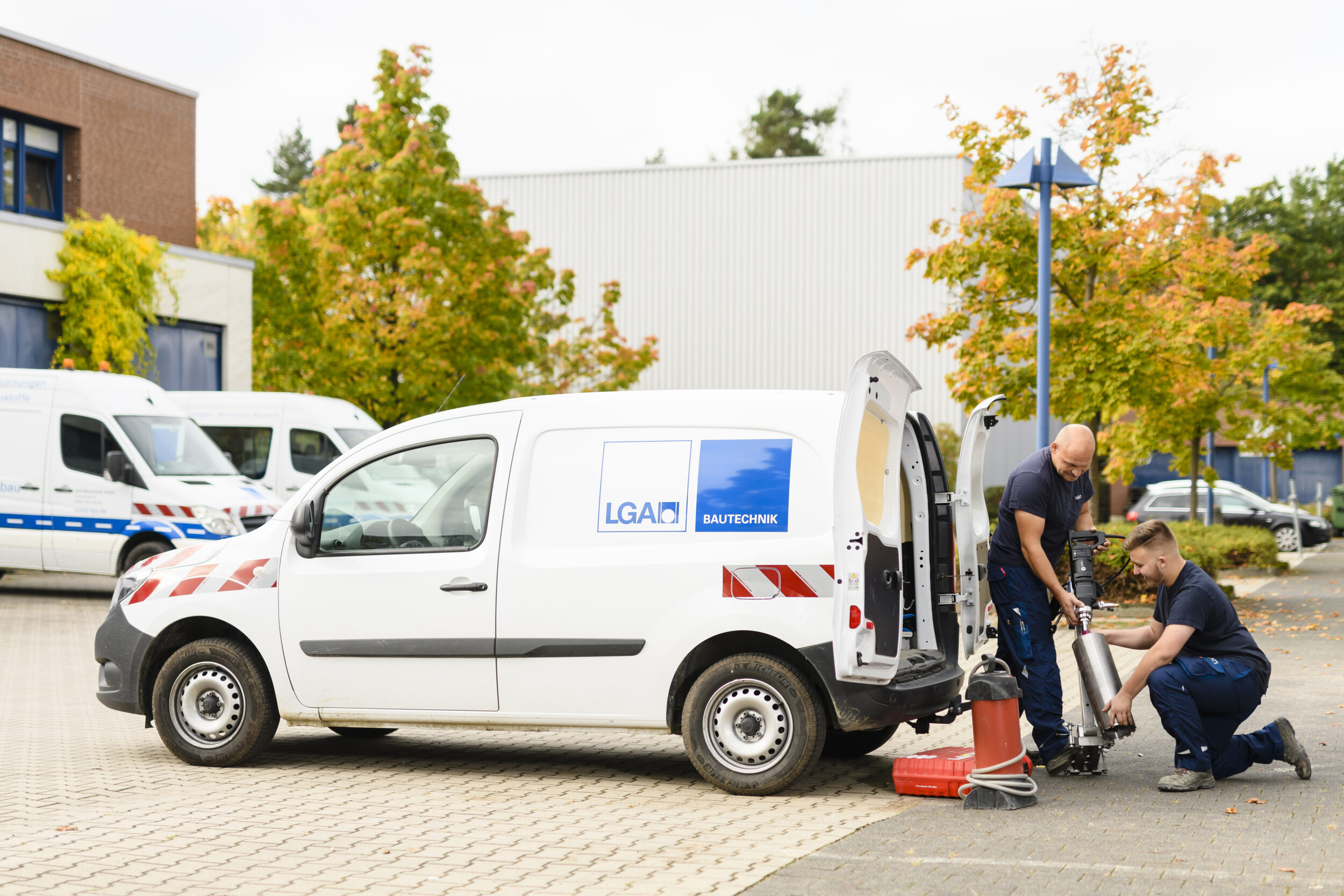 Das Bild zeigt zwei Personen bei der Arbeit neben einem weißen Transporter mit der Aufschrift „LGA BAUTECHNIK“. Die Hecktüren des Fahrzeugs sind geöffnet, und es sind verschiedene Geräte im und um den Wagen zu sehen. Im Hintergrund stehen weitere Fahrzeuge und Gebäude, umgeben von herbstlich gefärbten Bäumen.