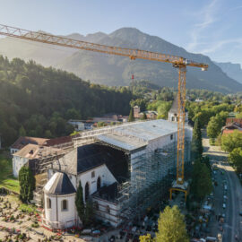 Das Bild zeigt eine große Baustelle, auf der eine Kirche renoviert wird. Die Kirche ist vollständig von Gerüsten umgeben, und zwei hohe Baukräne stehen links und rechts daneben. Im Hintergrund sind bewaldete Berge und verstreute Gebäude zu sehen, bei klarem Himmel mit wenigen Wolken.