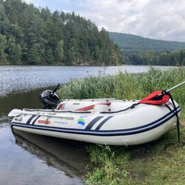 Ein aufblasbares Boot mit Motor liegt teils im Wasser, teils am Ufer eines Sees oder Flusses. Im Hintergrund sind bewaldete Uferbereiche und sanfte Hügel zu sehen.