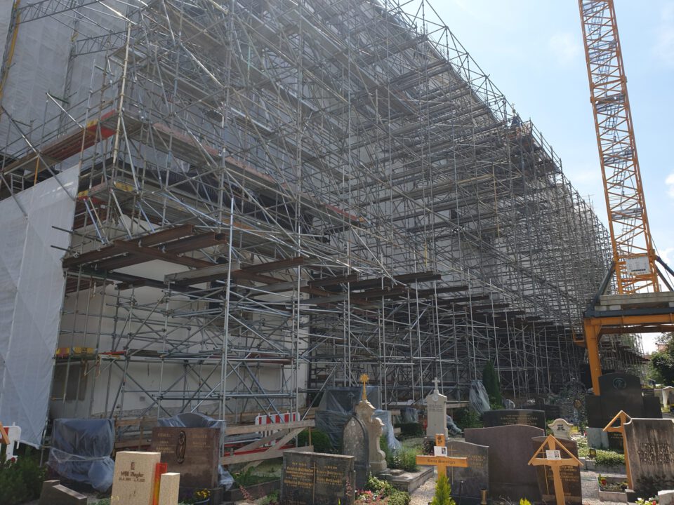 Das Bild zeigt eine große Baustelle mit umfangreichem Gerüst, das die gesamte Seite eines Gebäudes bedeckt. Rechts im Bild steht ein Baukran, und im Vordergrund befinden sich mehrere Grabsteine und Gräber, was auf eine Lage innerhalb oder neben einem Friedhof hinweist.