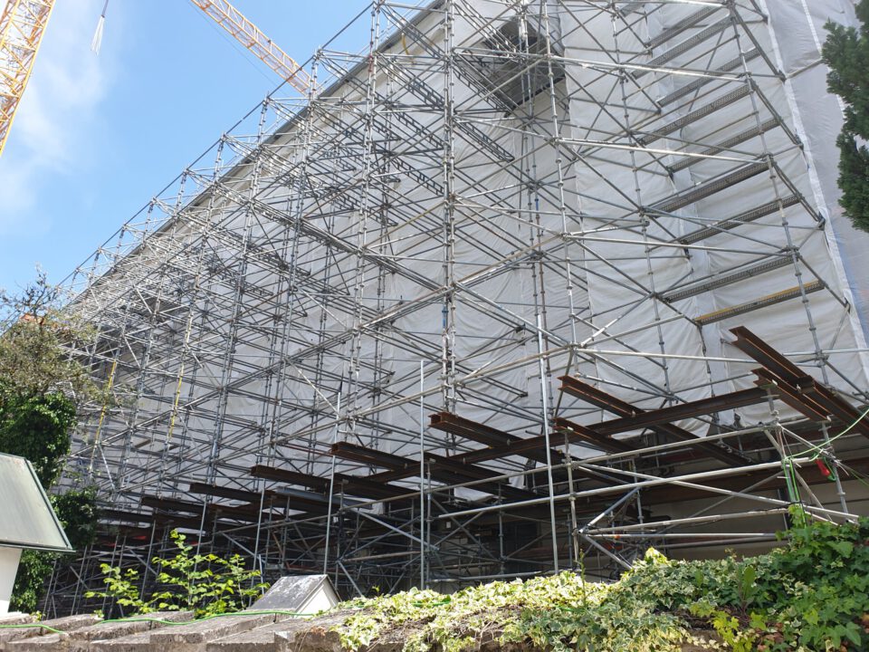 Das Bild zeigt eine große Baustelle mit umfangreichem Metallgerüst, das die sichtbare Seite eines Gebäudes vollständig umgibt und mit weißer Folie abgedeckt ist. Im Hintergrund sind Baukräne zu sehen, die auf laufende Arbeiten hinweisen. Im Vordergrund befinden sich Pflanzen und ein kleines Gebäude mit schrägem Dach.
