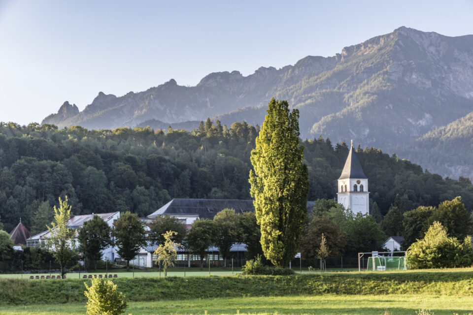 Das Bild zeigt eine idyllische Landschaft mit einem hohen Baum im Vordergrund und einer Kirche mit weißem Turm auf der rechten Seite. Mehrere Gebäude sind von Bäumen umgeben, im Hintergrund erstrecken sich bewaldete Hügel und Berge unter klarem Himmel. Eine offene Wiese liegt im Vordergrund.