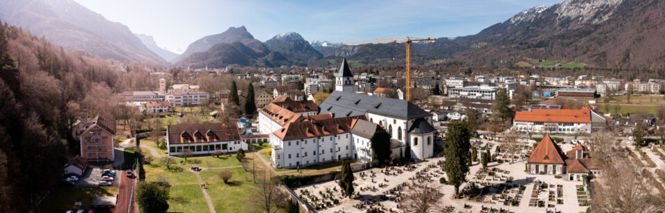 Das Bild zeigt eine Panoramaaufnahme eines Ortes inmitten einer Berglandschaft mit mehreren Gebäuden, darunter eine große Kirche mit hohem Turm und ein angrenzender Friedhof. Ein Baukran steht in der Nähe der Kirche, und im Hintergrund sind bewaldete Hügel sowie schneebedeckte Berge unter blauem Himmel zu sehen. Die Umgebung ist grün und gepflegt mit Wegen und Rasenflächen.