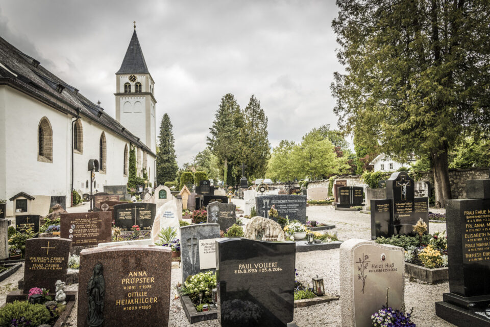 Das Bild zeigt eine Kirche mit hohem Glockenturm auf der linken Seite, umgeben von einem Friedhof. Die Kirche hat weiße Wände und Rundbogenfenster, und im Friedhof stehen zahlreiche Grabsteine und Denkmäler. Bäume und Grünflächen umrahmen die Szene, der Himmel ist wolkenverhangen.