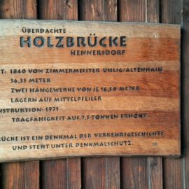 Auf dem Bild ist eine Informationstafel zur überdachten Holzbrücke Hennersdorf zu sehen. Es enthält Angaben zum Baujahr 1840, zur Länge der Brücke, zur Rekonstruktion im Jahr 1971 sowie zur Tragfähigkeit. Zudem wird darauf hingewiesen, dass die Brücke ein verkehrsgeschichtliches Denkmal ist und unter Denkmalschutz steht.