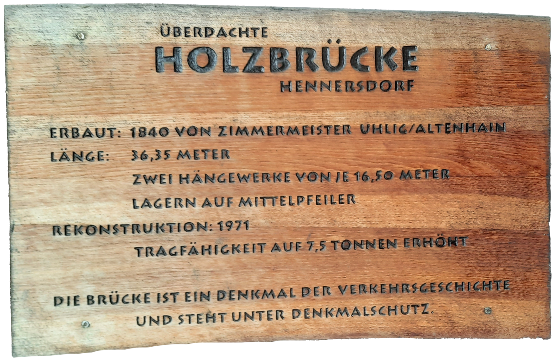 Auf dem Bild ist eine Informationstafel zur überdachten Holzbrücke Hennersdorf zu sehen. Es enthält Angaben zum Baujahr 1840, zur Länge der Brücke, zur Rekonstruktion im Jahr 1971 sowie zur Tragfähigkeit. Zudem wird darauf hingewiesen, dass die Brücke ein verkehrsgeschichtliches Denkmal ist und unter Denkmalschutz steht.