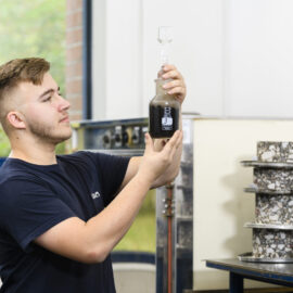 Das Bild zeigt eine Person in einem Labor, die ein Glasgefäß mit dunkler Flüssigkeit hochhält. Die Person trägt ein dunkelblaues Shirt. Im Hintergrund sind Laborgeräte und zylindrische Proben mit sichtbarem Gesteinsmaterial zu sehen.