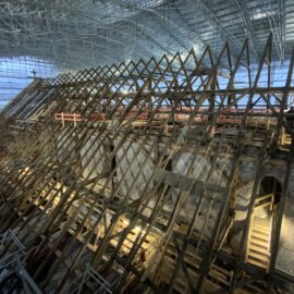 Das Bild zeigt eine große Baustelle im Inneren eines Gebäudes mit einem hölzernen Dachstuhl in Gitterstruktur. Um die Konstruktion herum befinden sich Gerüste und Arbeitsplattformen, was auf laufende Restaurierungsarbeiten hinweist. Im Hintergrund ist eine hohe Decke mit Metallträgern und Beleuchtung zu sehen, was auf ein historisches Gebäude wie eine Kathedrale schließen lässt.