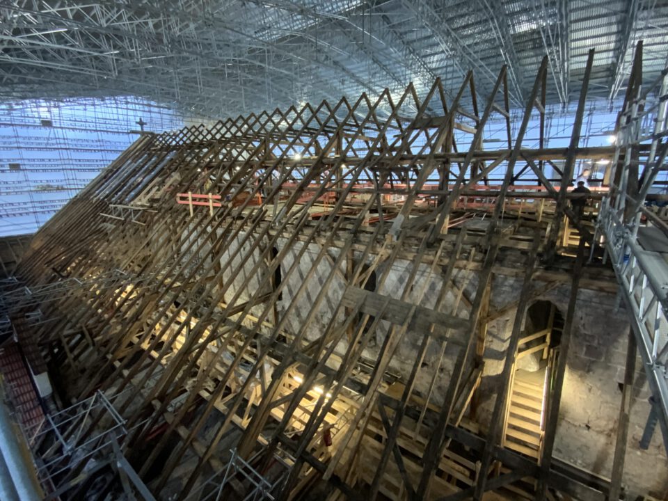 Das Bild zeigt eine große Baustelle im Inneren eines Gebäudes mit einem hölzernen Dachstuhl in Gitterstruktur. Um die Konstruktion herum befinden sich Gerüste und Arbeitsplattformen, was auf laufende Restaurierungsarbeiten hinweist. Im Hintergrund ist eine hohe Decke mit Metallträgern und Beleuchtung zu sehen, was auf ein historisches Gebäude wie eine Kathedrale schließen lässt.