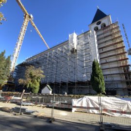 Das Bild zeigt ein großes Gebäude, vermutlich eine Kirche, das sich in Renovierung befindet und vollständig mit Gerüsten und Schutzplanen versehen ist. Links im Bild steht ein hoher Baukran, rechts ist ein Uhrturm mit spitzem Dach zu sehen. Vor dem Gebäude liegen Baumaterialien und Absperrungen, umgeben von Bäumen bei klarem, blauem Himmel.