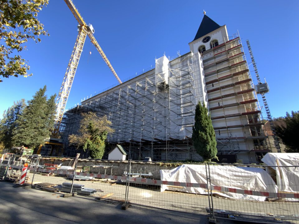 Das Bild zeigt ein großes Gebäude, vermutlich eine Kirche, das sich in Renovierung befindet und vollständig mit Gerüsten und Schutzplanen versehen ist. Links im Bild steht ein hoher Baukran, rechts ist ein Uhrturm mit spitzem Dach zu sehen. Vor dem Gebäude liegen Baumaterialien und Absperrungen, umgeben von Bäumen bei klarem, blauem Himmel.