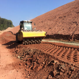 Das Bild zeigt eine Baustelle mit einem Bodenverdichter, der den Boden ebnet und verdichtet. Das Fahrzeug besitzt große Walzen mit Spitzen, die Abdrücke im Erdreich hinterlassen. Die Umgebung besteht aus roter Erde, rechts verläuft ein Hang, und im Hintergrund sind Bäume sowie weitere Fahrzeuge zu erkennen.