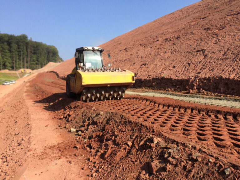 Das Bild zeigt eine Baustelle mit einem Bodenverdichter, der den Boden ebnet und verdichtet. Das Fahrzeug besitzt große Walzen mit Spitzen, die Abdrücke im Erdreich hinterlassen. Die Umgebung besteht aus roter Erde, rechts verläuft ein Hang, und im Hintergrund sind Bäume sowie weitere Fahrzeuge zu erkennen.