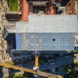 Das Bild zeigt eine Luftaufnahme eines großen Gebäudes, das sich im Bau oder in der Renovierung befindet und mit einer provisorischen Dachkonstruktion überdeckt ist. Ein gelber Baukran ragt vom unteren Bildrand in Richtung Gebäudemitte, umgeben von kleineren Gebäuden mit roten Dächern und einer Straße mit Fahrzeugen. Auf einer Seite des Gebäudes befindet sich ein Bereich mit vielen kleinen Strukturen in Reihen, vermutlich ein Friedhof oder Garten.