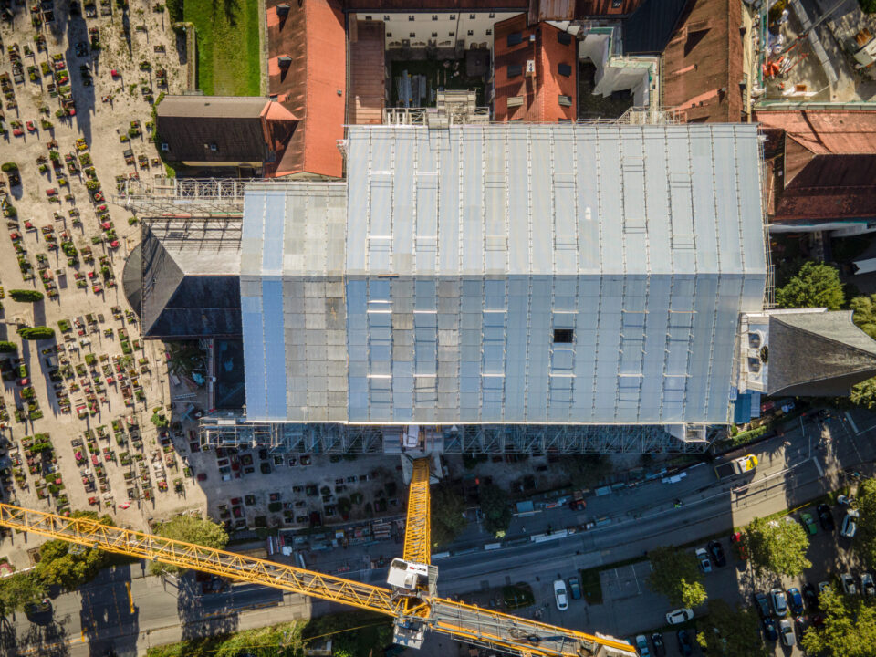 Das Bild zeigt eine Luftaufnahme eines großen Gebäudes, das sich im Bau oder in der Renovierung befindet und mit einer provisorischen Dachkonstruktion überdeckt ist. Ein gelber Baukran ragt vom unteren Bildrand in Richtung Gebäudemitte, umgeben von kleineren Gebäuden mit roten Dächern und einer Straße mit Fahrzeugen. Auf einer Seite des Gebäudes befindet sich ein Bereich mit vielen kleinen Strukturen in Reihen, vermutlich ein Friedhof oder Garten.