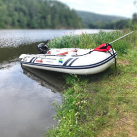Ein aufblasbares Boot mit Motor liegt teils im Wasser, teils am Ufer eines Sees oder Flusses. Im Hintergrund sind bewaldete Uferbereiche und sanfte Hügel zu sehen.