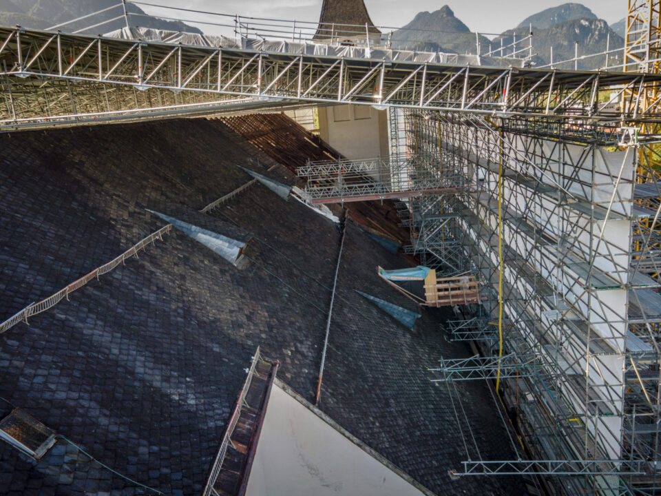 Das Bild zeigt eine Baustelle auf dem Dach eines großen Gebäudes, das mit dunklen Schindeln gedeckt ist. Um das Dach herum sind mehrere Gerüstkonstruktionen aufgebaut, und eine provisorische Brücke aus Metallträgern verläuft quer über das Dach. Im Hintergrund sind Berge unter klarem Himmel zu sehen.