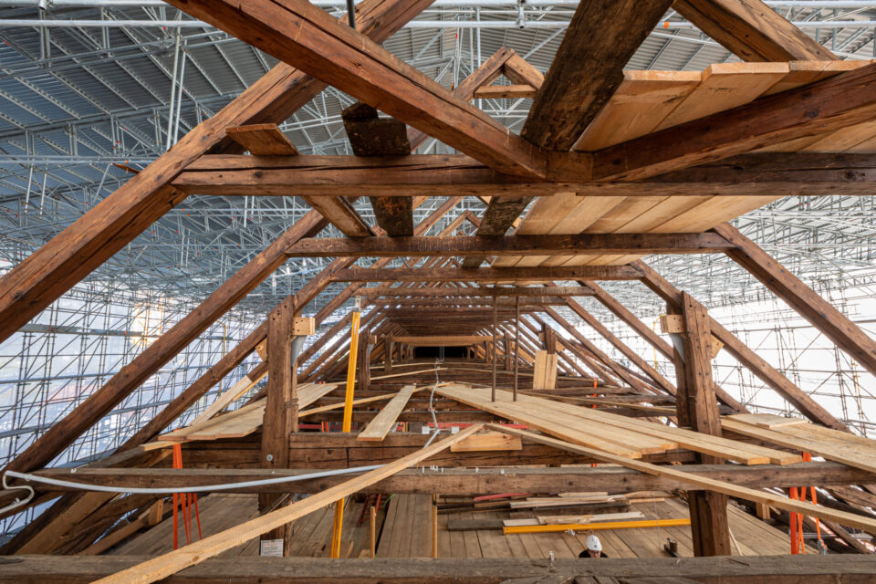 Das Bild zeigt das Innere einer großen Holzkonstruktion, die sich im Bau oder in der Renovierung befindet. Der Blick verläuft durch einen langen Gang aus hölzernen Trägern und Planken, die in einem dreieckigen Muster das Dach stützen. Im Hintergrund sind Gerüste und Baumaterialien wie Holzplanken und Metallstangen zu sehen.