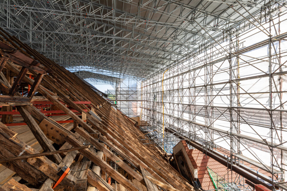 Das Bild zeigt eine große Baustelle im Inneren eines Gebäudes mit umfangreichem Gerüst auf der rechten Seite, bestehend aus Metallträgern in Gitterstruktur. Links befindet sich eine geneigte Holzkonstruktion, vermutlich Teil eines Dachs oder Bodens in Bau oder Sanierung. Eine provisorische Überdachung aus Metall schützt die Baustelle vor Witterungseinflüssen.