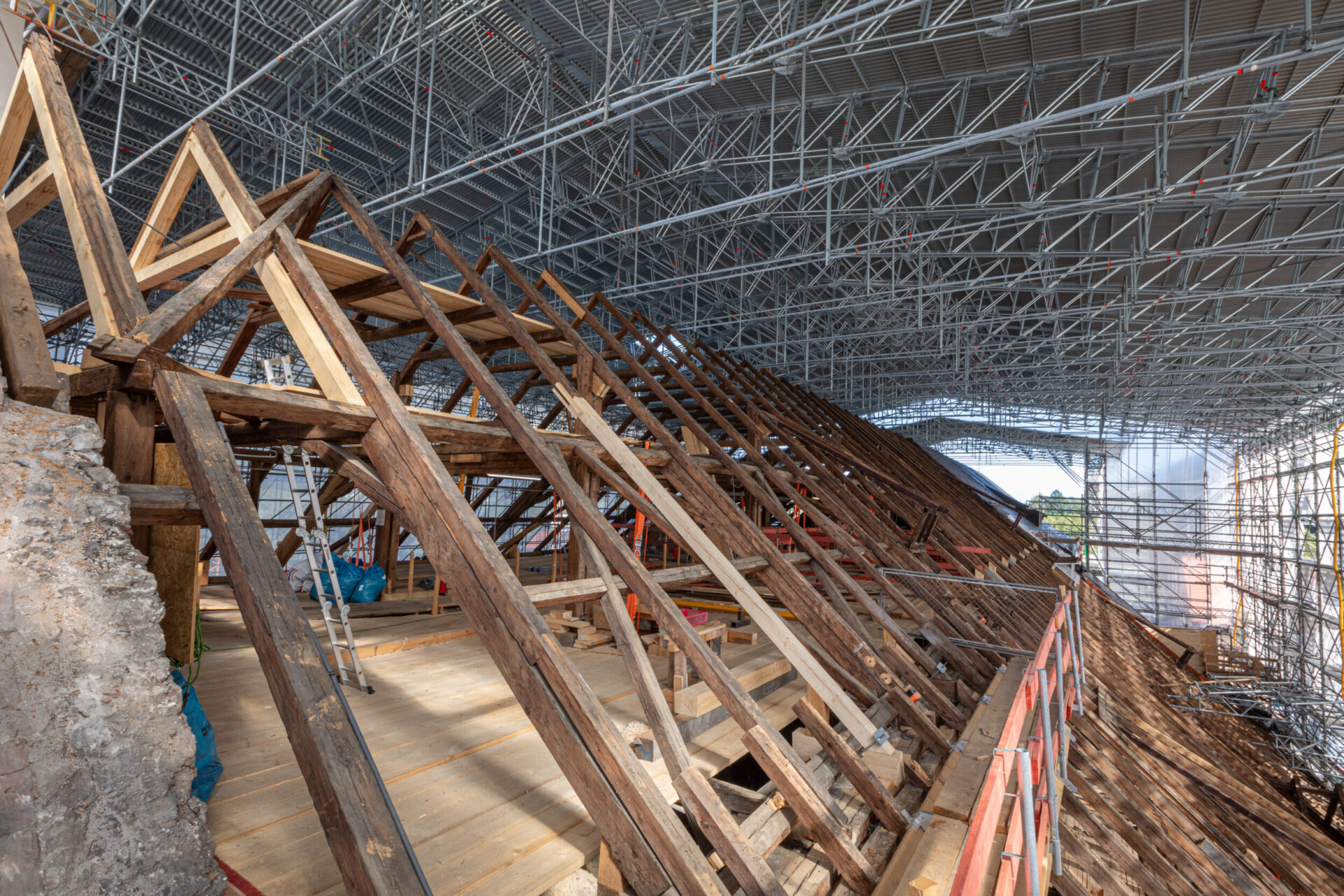 Das Bild zeigt eine große Baustelle im Inneren eines Gebäudes mit umfangreichem Metallgerüst und einem hölzernen Dachstuhl aus mehreren dreieckigen Trägern und Balken. Leitern und Baumaterialien deuten auf laufende Arbeiten hin. Eine schützende Metallüberdachung überspannt den gesamten Bereich, was auf die Sanierung eines historischen Gebäudes schließen lässt.
