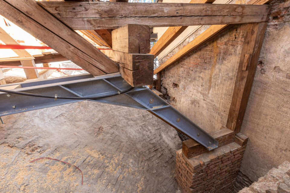 Das Bild zeigt eine große Baustelle im Inneren eines Gebäudes mit umfangreichem Metallgerüst und einem hölzernen Dachstuhl aus mehreren dreieckigen Trägern und Balken. Leitern und Baumaterialien deuten auf laufende Arbeiten hin. Eine schützende Metallüberdachung überspannt den gesamten Bereich, was auf die Sanierung eines historischen Gebäudes schließen lässt.